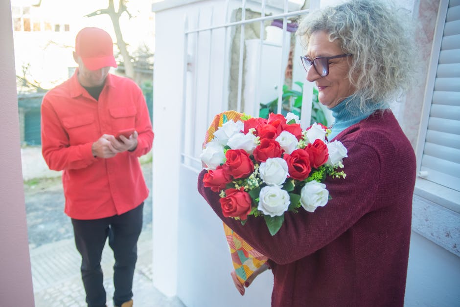 A woman with curly gray hair and glasses holding a vibrant floral bouquet featuring red and white roses, wrapped in orange and yellow patterned paper, during a flower delivery at her doorstep. The wom