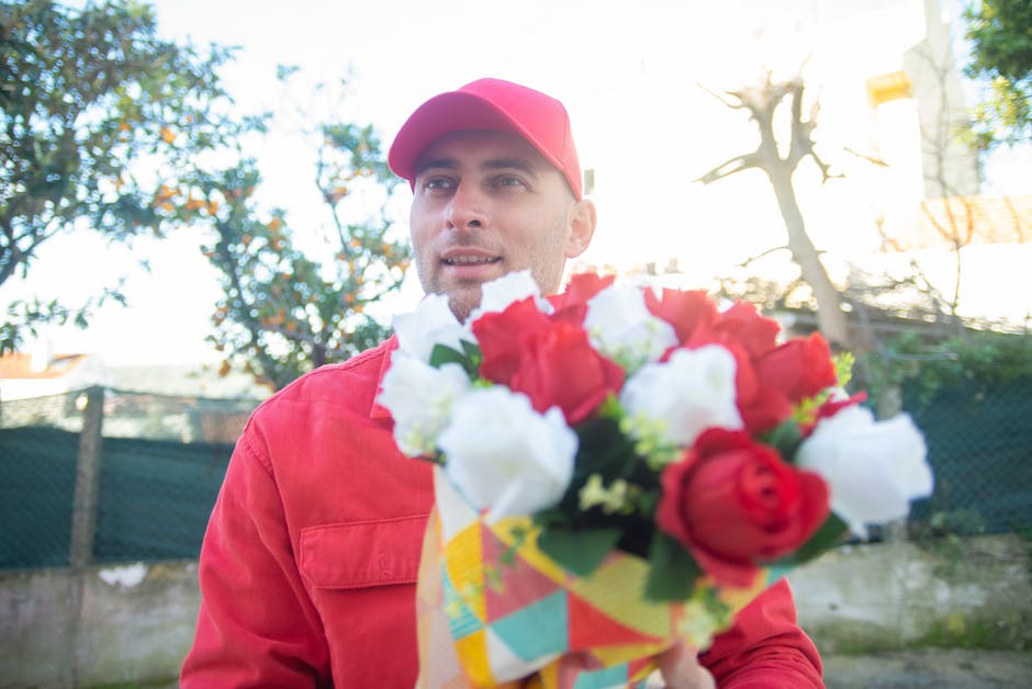 A delivery person dressed in a red uniform and matching cap is holding a vibrant floral bouquet outdoors, with a concrete fence, green hedge, and trees in the background. The bouquet features a mix of