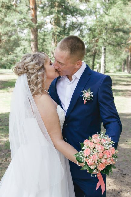 A newlywed couple shares a romantic kiss outdoors in a lush, green park with tall trees in the background. The bride has blonde, wavy hair and is wearing a traditional white wedding gown with a veil, 