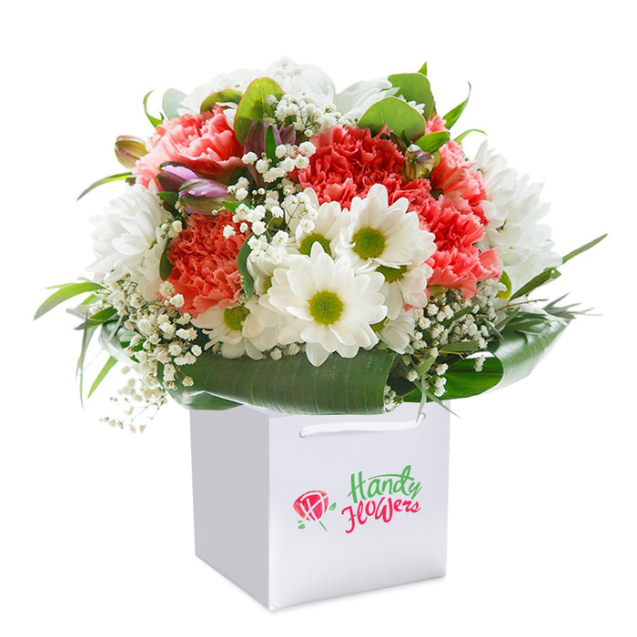 Against a flawless white backdrop, this expertly composed bouquet radiates the artistry and freshness that Marks Gate locals expect from their community florists. Perched upright in a clean, modern white gift bag and made ready for easy delivery across Marks Gate, the arrangement reveals a vibrant spectrum: from blushing coral and peach hues to deep, passionate red carnations, known for their elegantly ruffled petals. Brimming amongst them are snowy white daisies-each centered with a fresh, lime-tinted yellow-that infuse the bouquet with a playful, uplifting spirit. Tiny clusters of white baby's breath are delicately woven throughout, lending an ethereal, weightless quality to the dense, full shape. Close examination reveals buds with a faint wash of purple on the left side, adding intrigue and suggesting the perpetual renewal of gardens glimpsed along Lodge Avenue. At the base, broad, dark green leaves create a lush collar, their gleaming surfaces forming a vivid contrast to the soft petals above. The pristine white gift bag, secured with tastefully sturdy ribbon handles, remains free of exterior branding, keeping the flowers themselves as the focal point. Every element is arranged to perfection: a harmonious, rounded profile that speaks of freshness and care, destined for occasions such as anniversaries, local celebrations, or thoughtful surprises. The interplay of textures, from velvet petals to glossy greens, reflects the pride and expertise of a seasoned Marks Gate florist-someone who understands the area's unique fabric and is devoted to delivering beauty straight to neighbours' doorsteps.