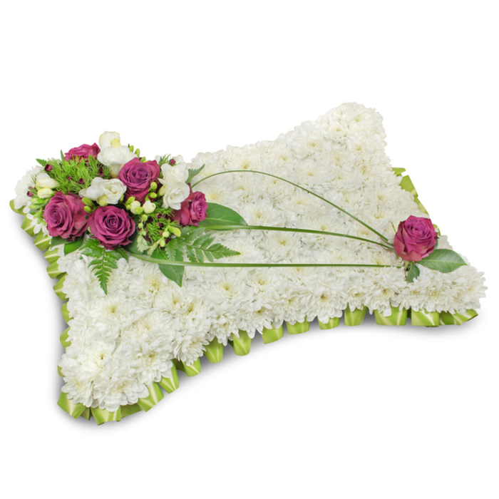 Set against a clean white background and resting on a faint shadow, this traditional pillow-shaped sympathy arrangement is a study in quiet craftsmanship and meaning. The surface is entirely covered with hundreds of small, pure white chrysanthemum florets-each one tightly formed-producing a plush, cloudlike field that symbolizes peace and purity. A continuous trim of folded lime-green satin ribbon encircles the pillow, its pleated edges creating a refined border that frames the white blooms with subtle colour and structure. Dominating the upper-left corner, a cluster of rich magenta roses opens in velvety layers, interspersed with slender white freesia and occasional lisianthus, offering a soft perfume and delicate, contrasting highlights. Underpinning the flowers is a generous selection of deep green foliage, including feathery fern fronds, which adds weight and depth and lifts the focal cluster. Three long, dark-green bear grass stems sweep diagonally across the chrysanthemum plane, their flowing lines introducing movement and visual balance; on the opposite side a single matching magenta rose is placed near the lower-right edge to complete the symmetry. The lighting is soft, neutral and even, bringing out the texture of the petals and the satin sheen of the ribbon. This arrangement, suitable for a funeral or memorial service in Marks Gate, Chadwell Heath and neighbouring Barking, carries the quiet dignity of a well-made tribute-an offering of comfort and remembrance from Flowers Marks Gate, crafted with local care and floral expertise.